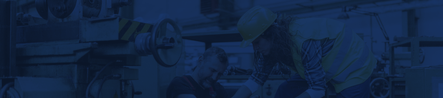Factory worker wearing a safety vest and hard hat assisting an injured coworker beside industrial machinery inside a workshop.