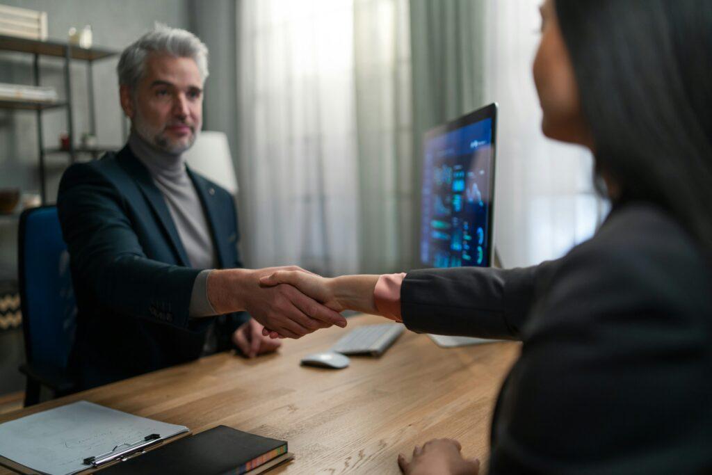 Two professionals in business attire shaking hands across a desk in an office, symbolizing agreement, trust, or the start of a legal consultation.