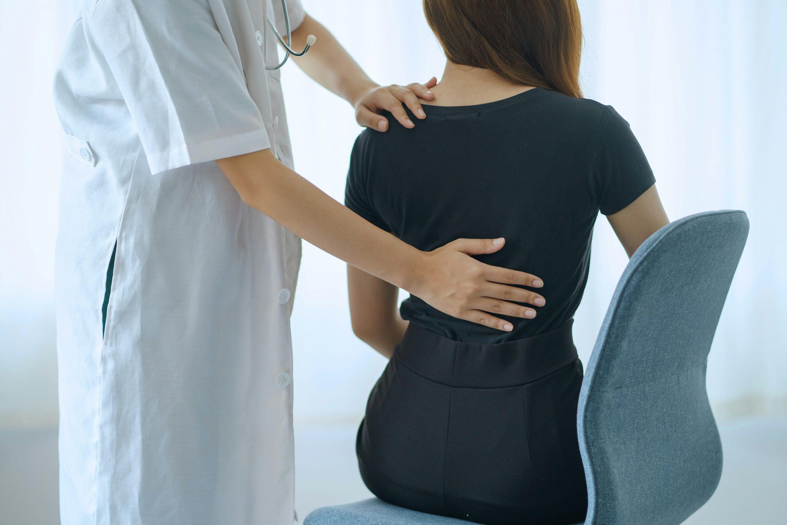 Doctor examining a patient’s back, placing hands on the upper and lower back areas while the patient sits on a chair during a medical consultation.