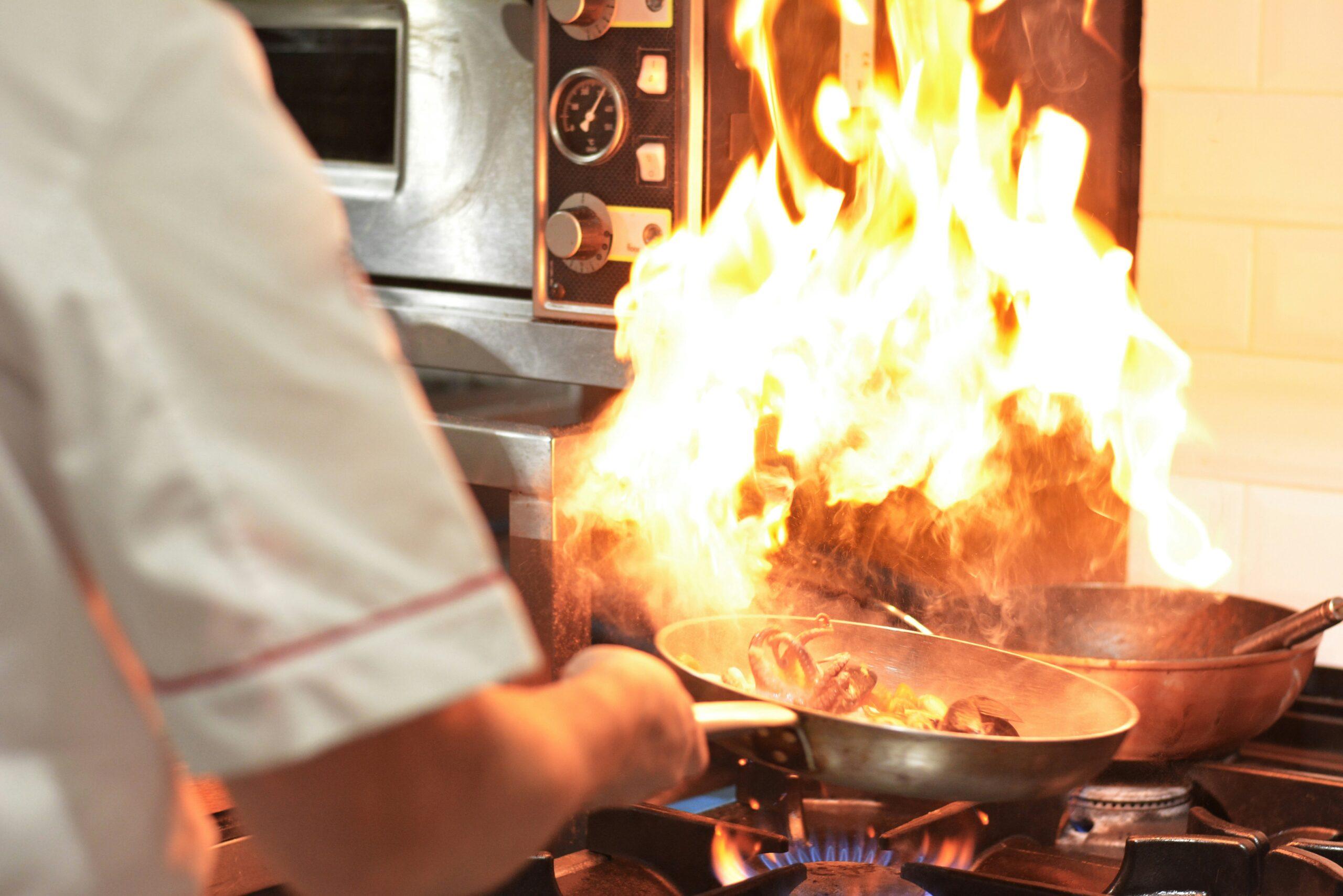 Chef cooking on a gas stove as flames flare up from a frying pan in a busy kitchen.