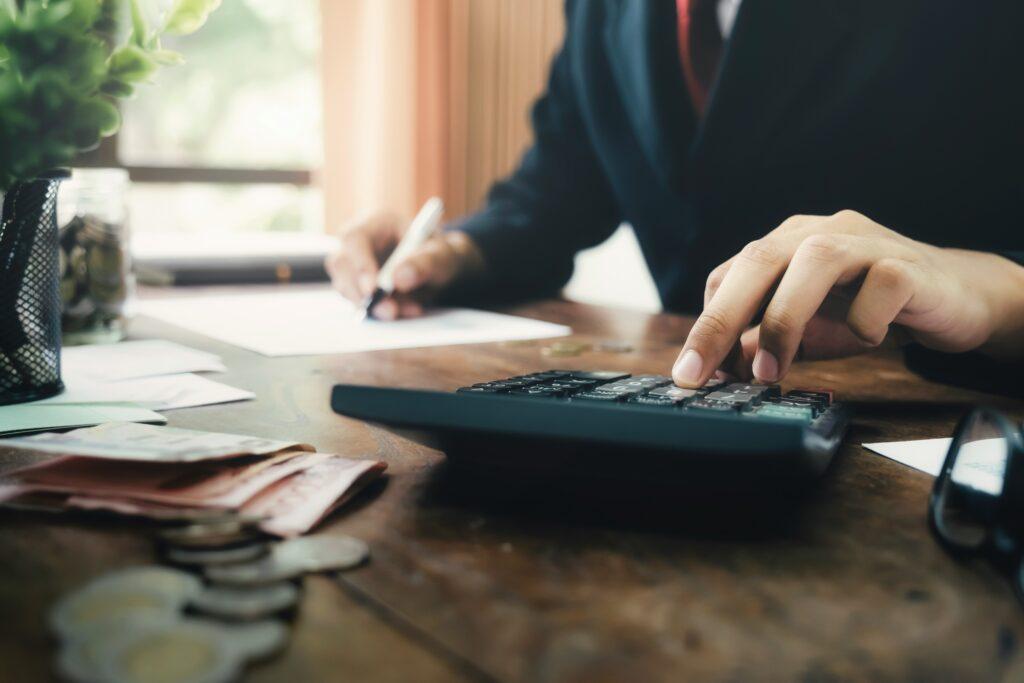 Person in a business suit using a calculator and writing on paper at a wooden desk with money, coins, and documents scattered around.