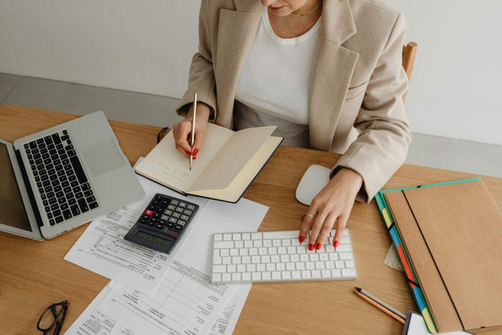 Woman working at a desk with a laptop, keyboard, calculator, and documents, writing notes in a notebook while organizing paperwork and folders.