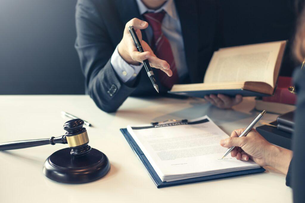 Lawyer consulting with a client at a desk, holding a pen and open law book while the client signs legal documents beside a judge’s gavel.