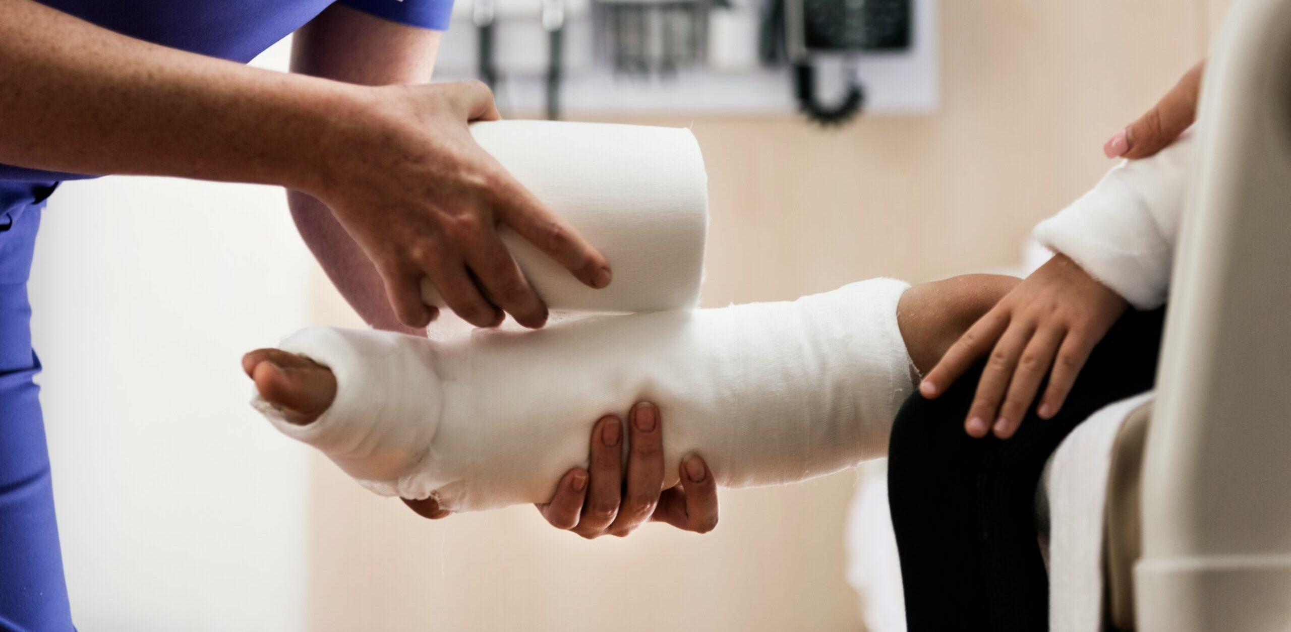 Healthcare professional applying a cast to a young patient’s leg while the child sits on a hospital bed with an already bandaged arm.