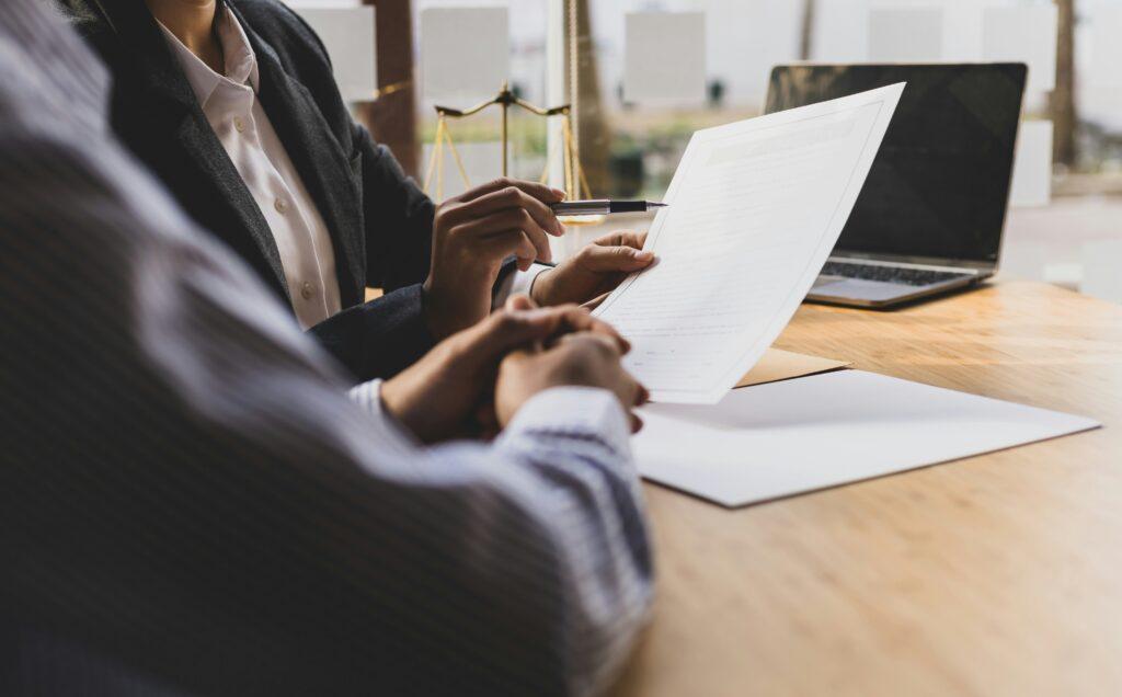 Lawyer reviewing legal documents with a client at a desk, with a laptop and scales of justice in the background.