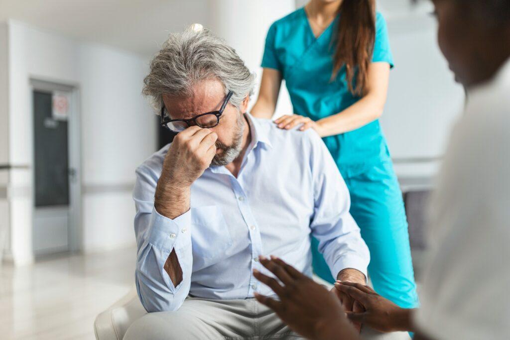 Elderly man sitting in a medical facility holding his forehead in distress while a nurse places a comforting hand on his shoulder during a consultation.