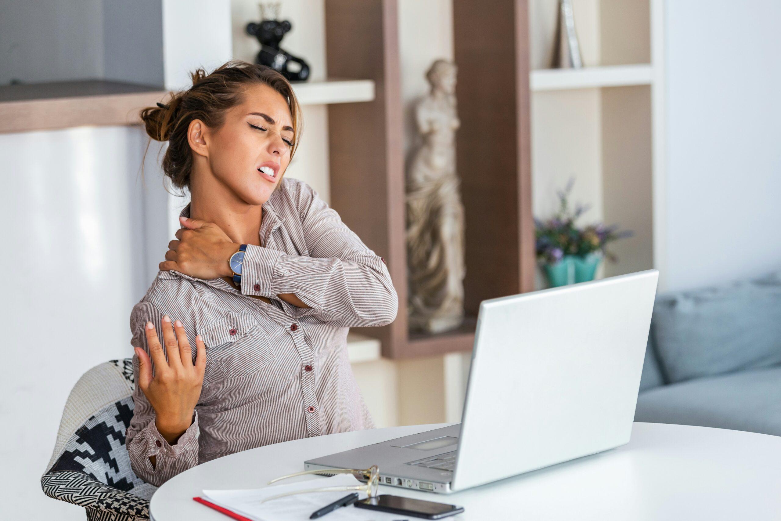 Woman sitting at a desk in front of a laptop, holding her neck and shoulder in pain, showing discomfort likely from muscle strain or poor posture while working on a computer.
