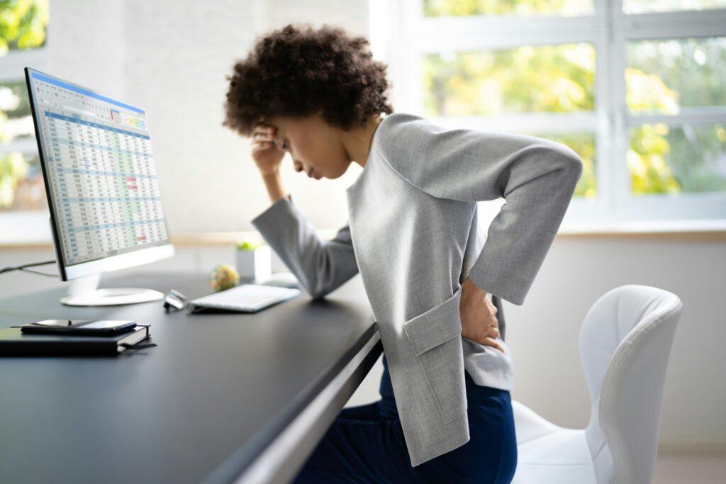 Woman sitting at an office desk in front of a computer, holding her lower back in pain and leaning forward, showing discomfort from back strain or poor posture.