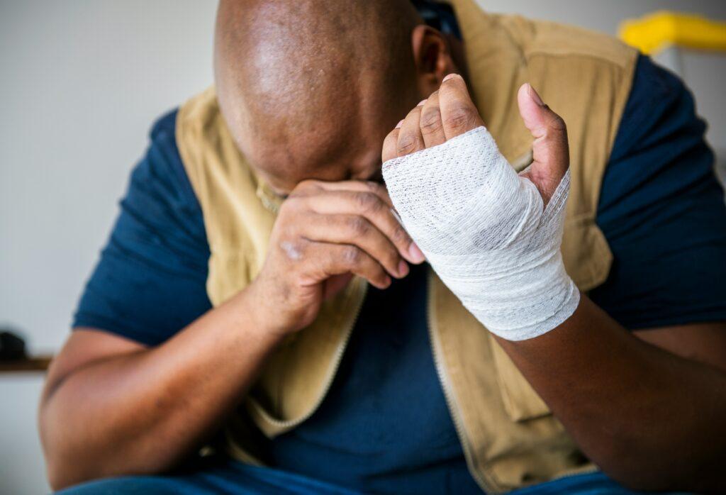 Man sitting with his head lowered, holding his bandaged hand in pain, showing signs of injury and emotional distress.
