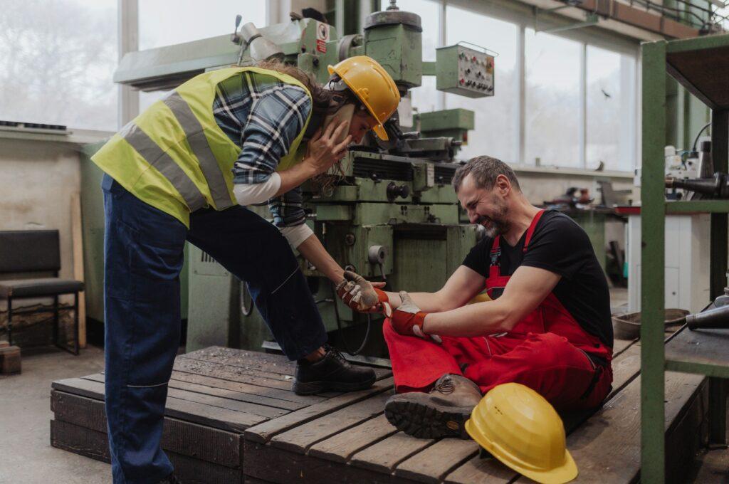 Injured factory worker sitting on the floor holding his leg in pain while a coworker in a safety vest and helmet calls for help inside an industrial workshop.