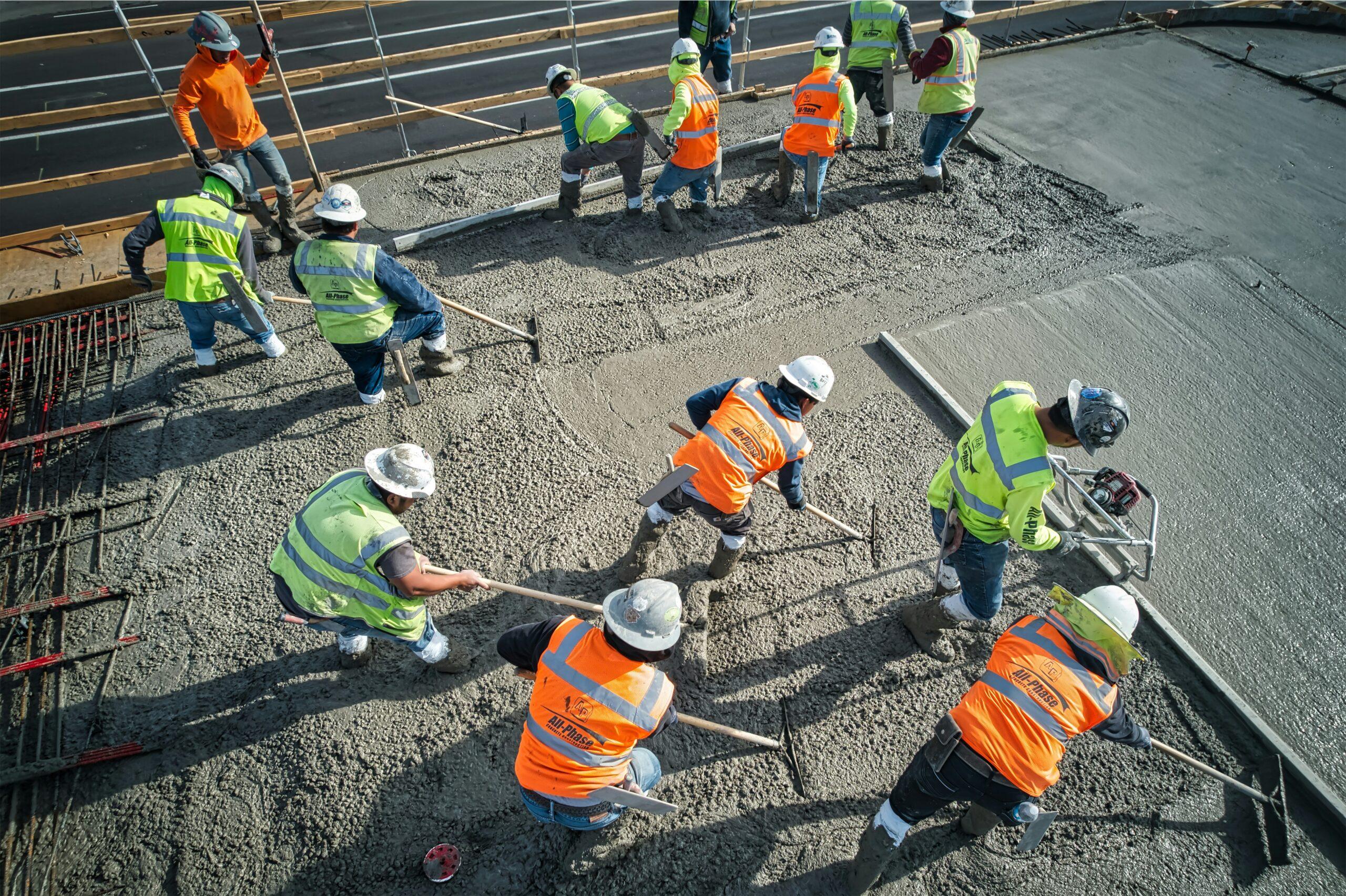 Construction workers in bright safety vests and helmets smoothing and leveling wet concrete on a large job site, using long-handled tools while standing in the freshly poured surface.