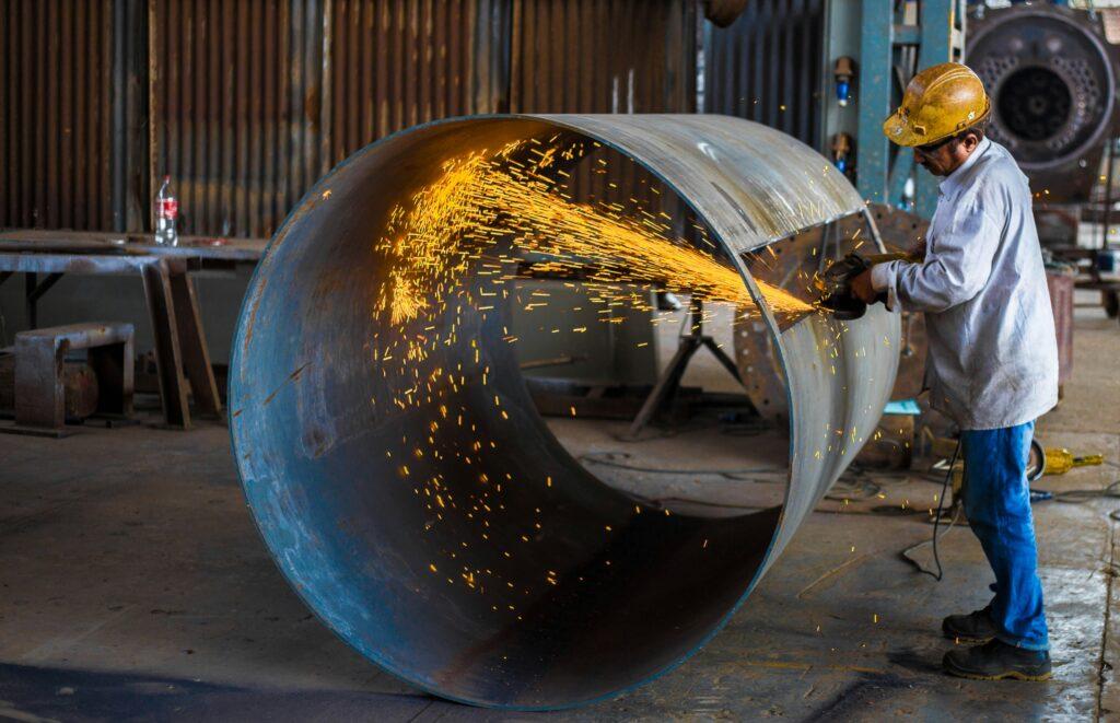 A worker in protective gear uses a grinder to cut through a large metal pipe, sending bright sparks flying across an industrial workshop.