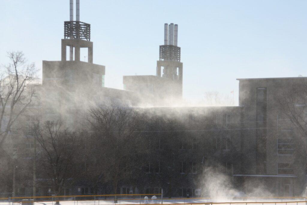 An industrial or institutional building with multiple rooftop stacks releasing visible steam or vapor into cold air, bare winter trees in the foreground, and a pale blue sky creating a hazy, wintry atmosphere around the structure.
