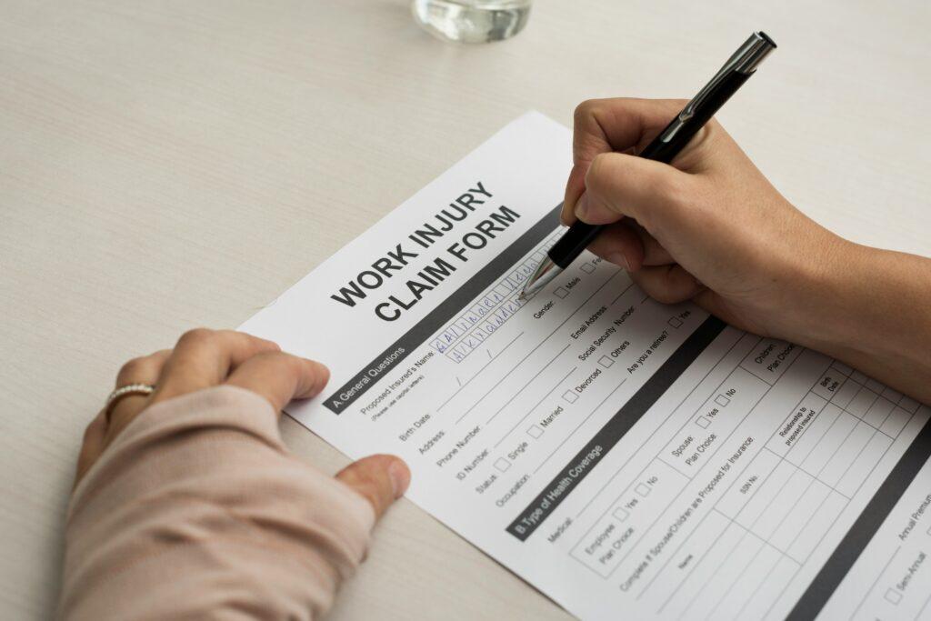 A close-up view of someone filling out a work injury claim form at a desk, with one hand writing in the personal-information fields using a black pen while the other hand holds the form steady.
