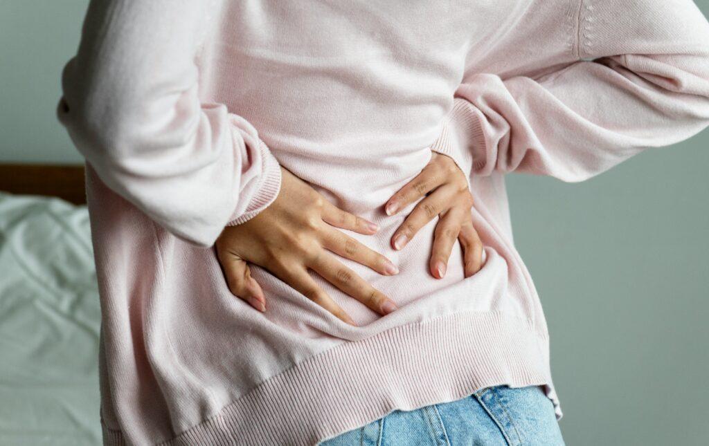 A person wearing a light pink sweater and jeans holding their lower back with both hands, slightly bent forward as if experiencing sharp lumbar pain.