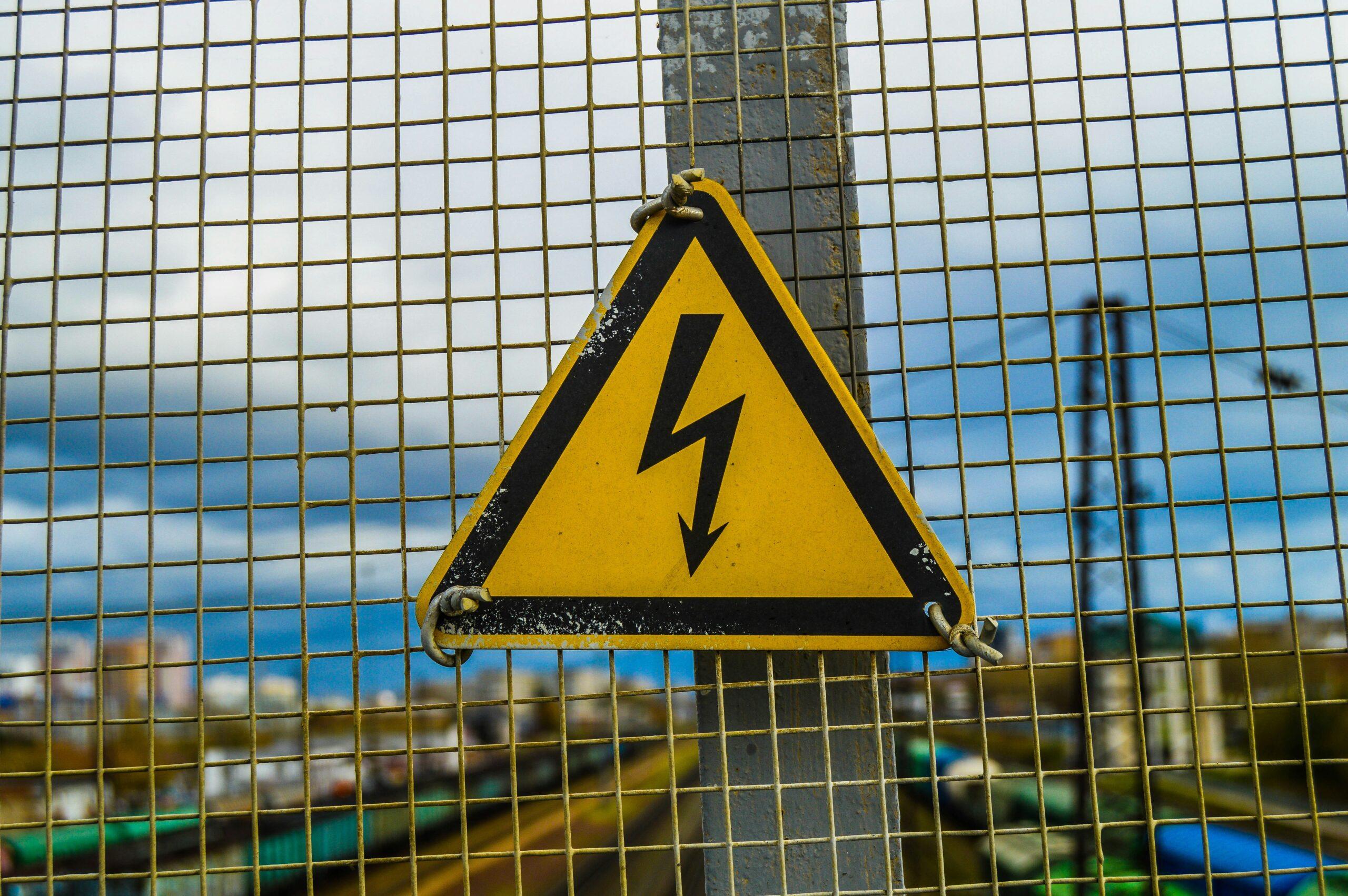A worn yellow high-voltage warning sign with a black lightning-bolt symbol hangs on a metal fence above what appears to be a railway or industrial area, with blurred tracks and buildings in the background under a cloudy sky.
