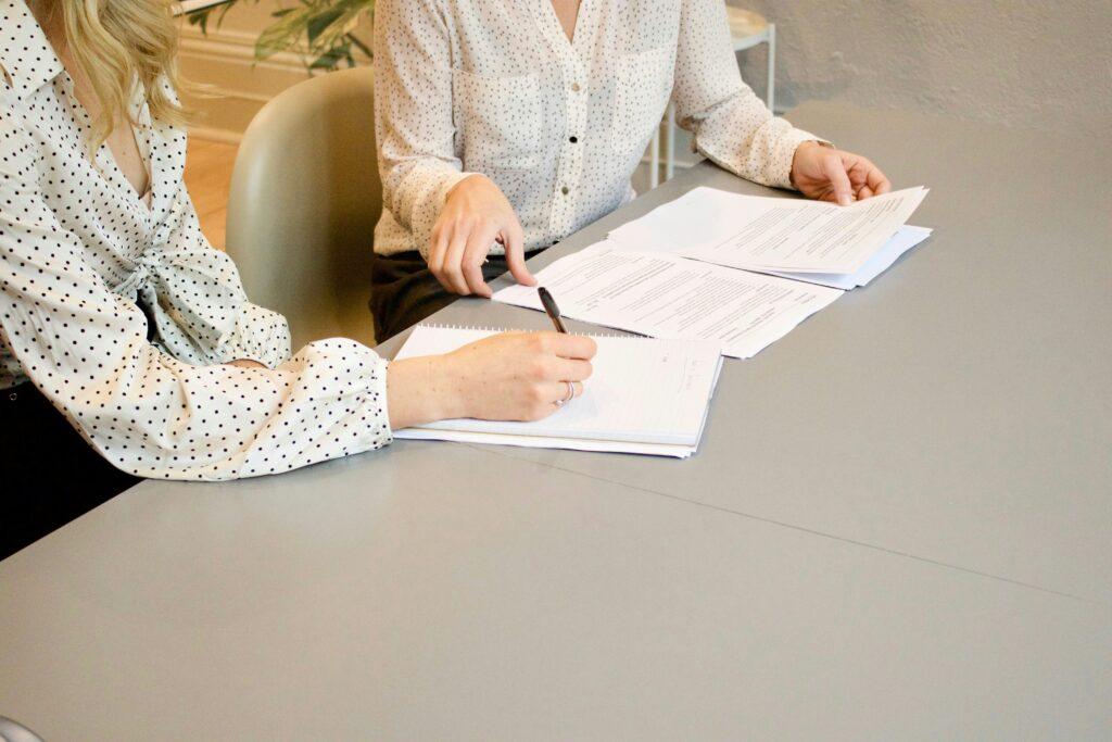 Two people in light, patterned blouses sitting at a table reviewing documents, one taking notes in a notebook while the other points toward a stack of printed papers.
