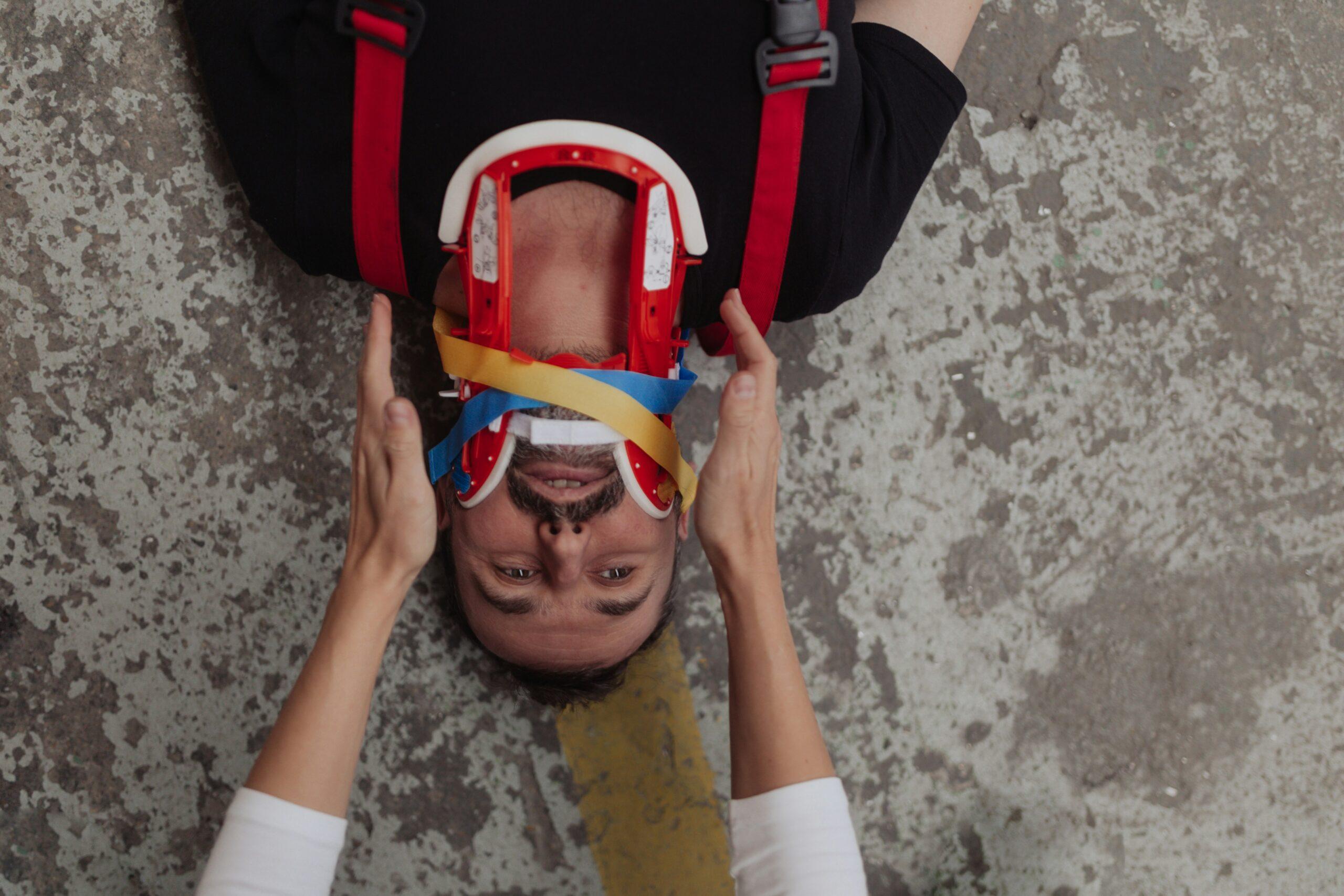 A person lies on the ground restrained in a red-and-white cervical immobilization collar, with colorful medical straps securing their head while another individual carefully stabilizes it with both hands.