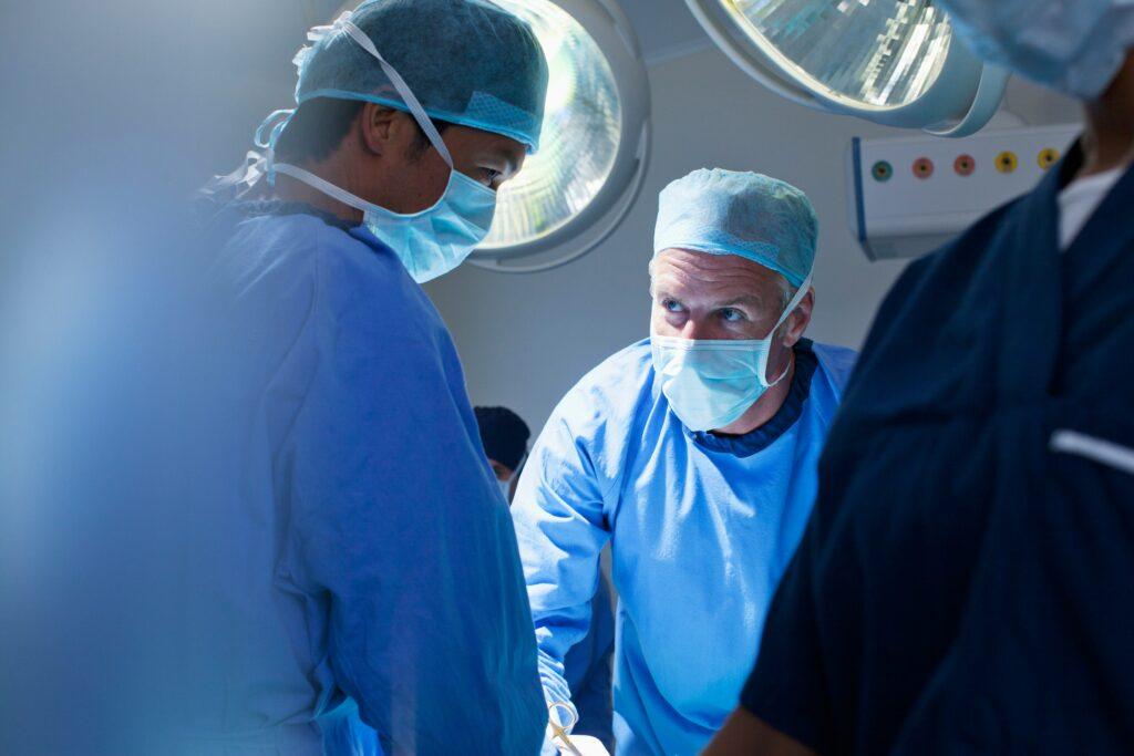 A team of surgeons in full protective gear working under bright operating room lights, with one surgeon intently focused while speaking to a colleague during a procedure.
