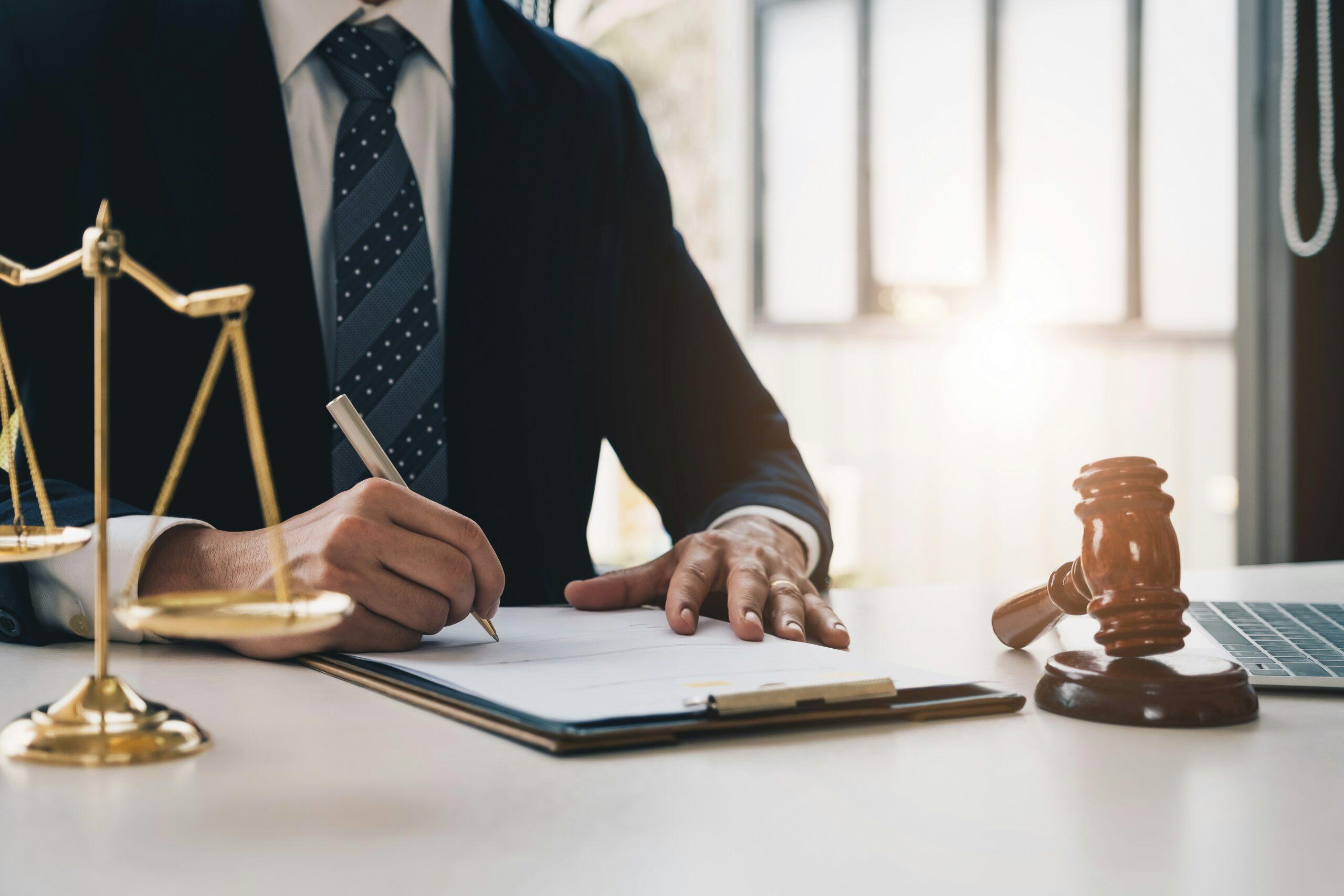 A lawyer in a suit writing on documents at a desk, with a gavel and scales of justice nearby in a sunlit office.