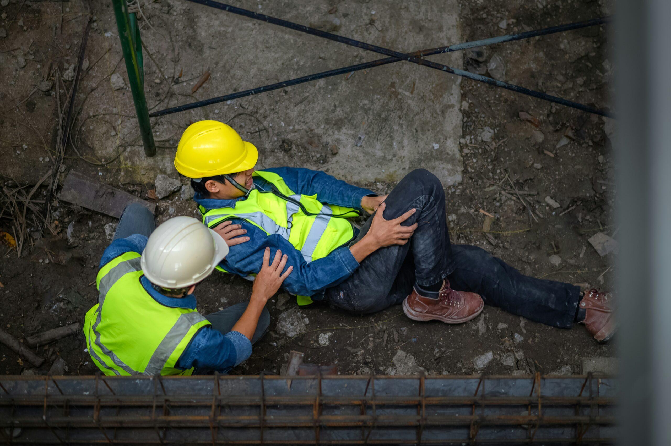A construction worker wearing a yellow hard hat and safety vest lies on the ground clutching his leg, while a coworker in similar safety gear kneels beside him offering support at the accident site.