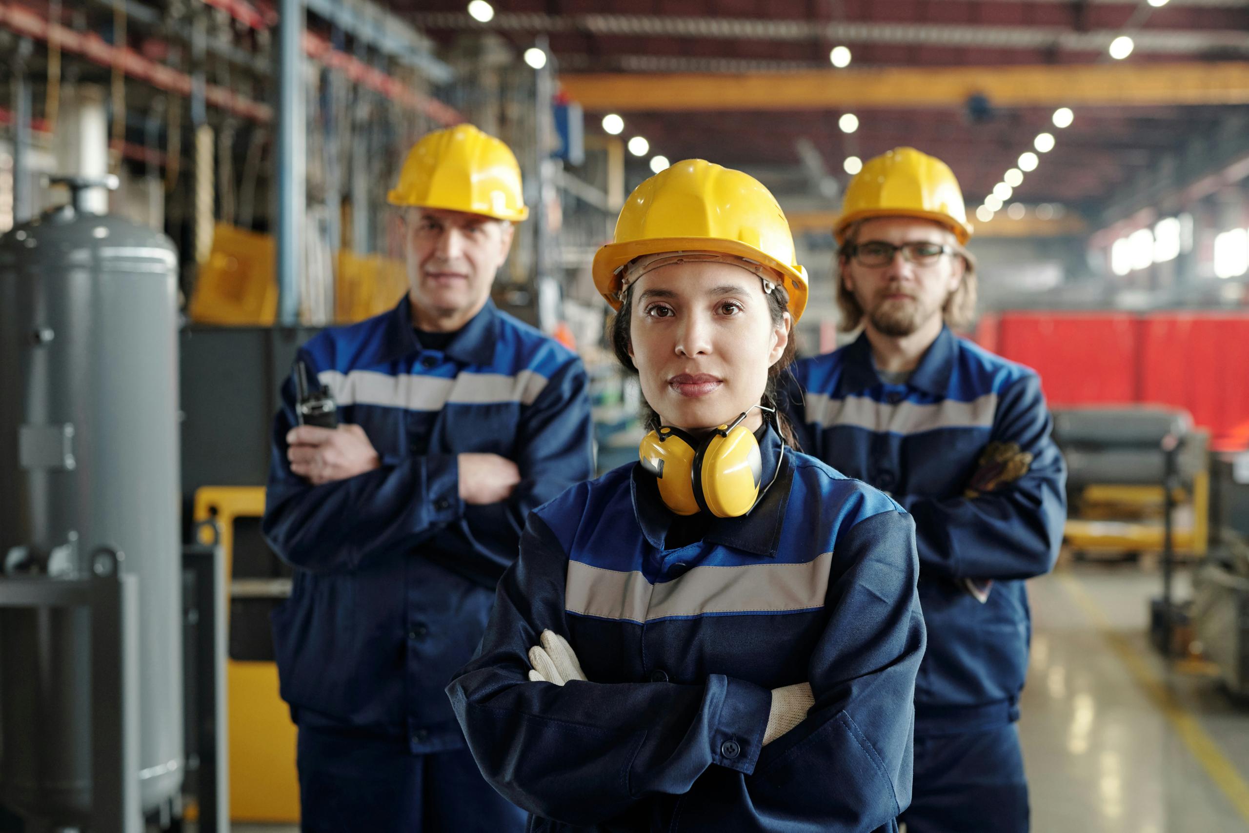 A group of industrial workers in protective gear standing confidently in a factory, wearing yellow hard hats, blue uniforms, and safety equipment.