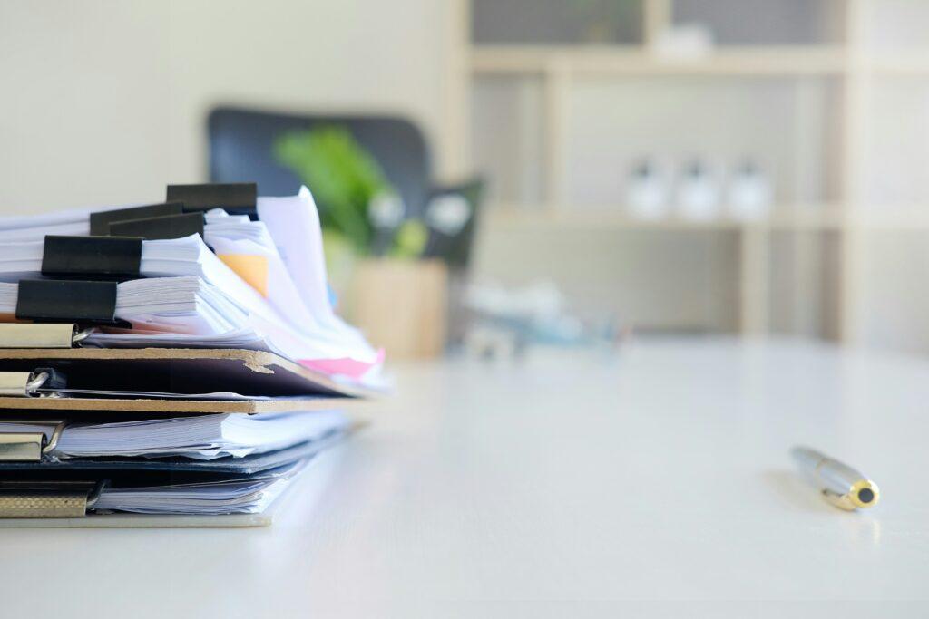 A stack of overloaded folders and clipped documents sits on a bright office desk, with papers slightly sticking out and edges worn, while a pen rests on the otherwise clear surface and blurred shelves and plants appear in the background.