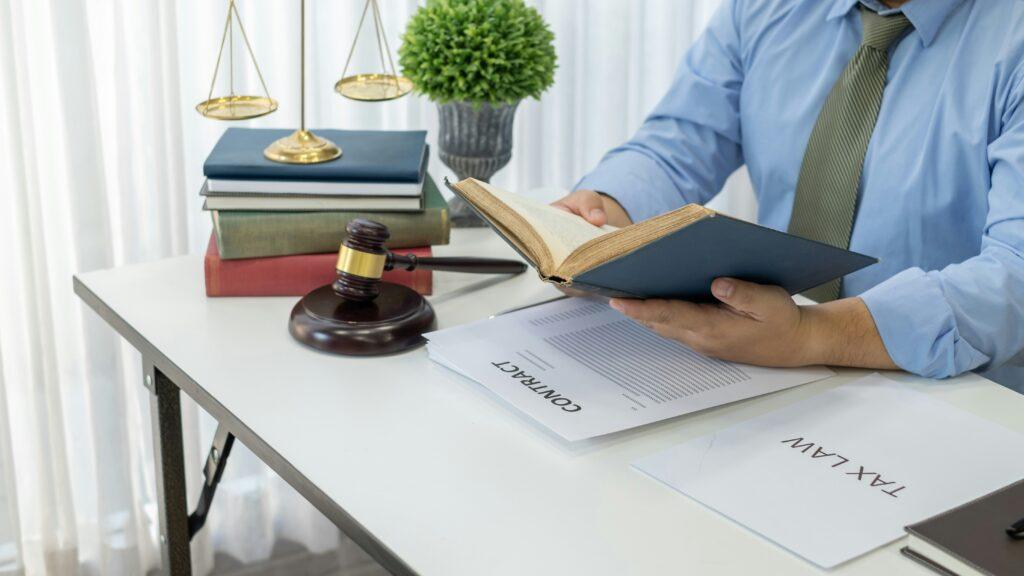 Person in a blue shirt and tie reading an open law book at a white desk with a wooden gavel, stacked legal books, balance scales, and printed contract and tax law documents arranged neatly nearby