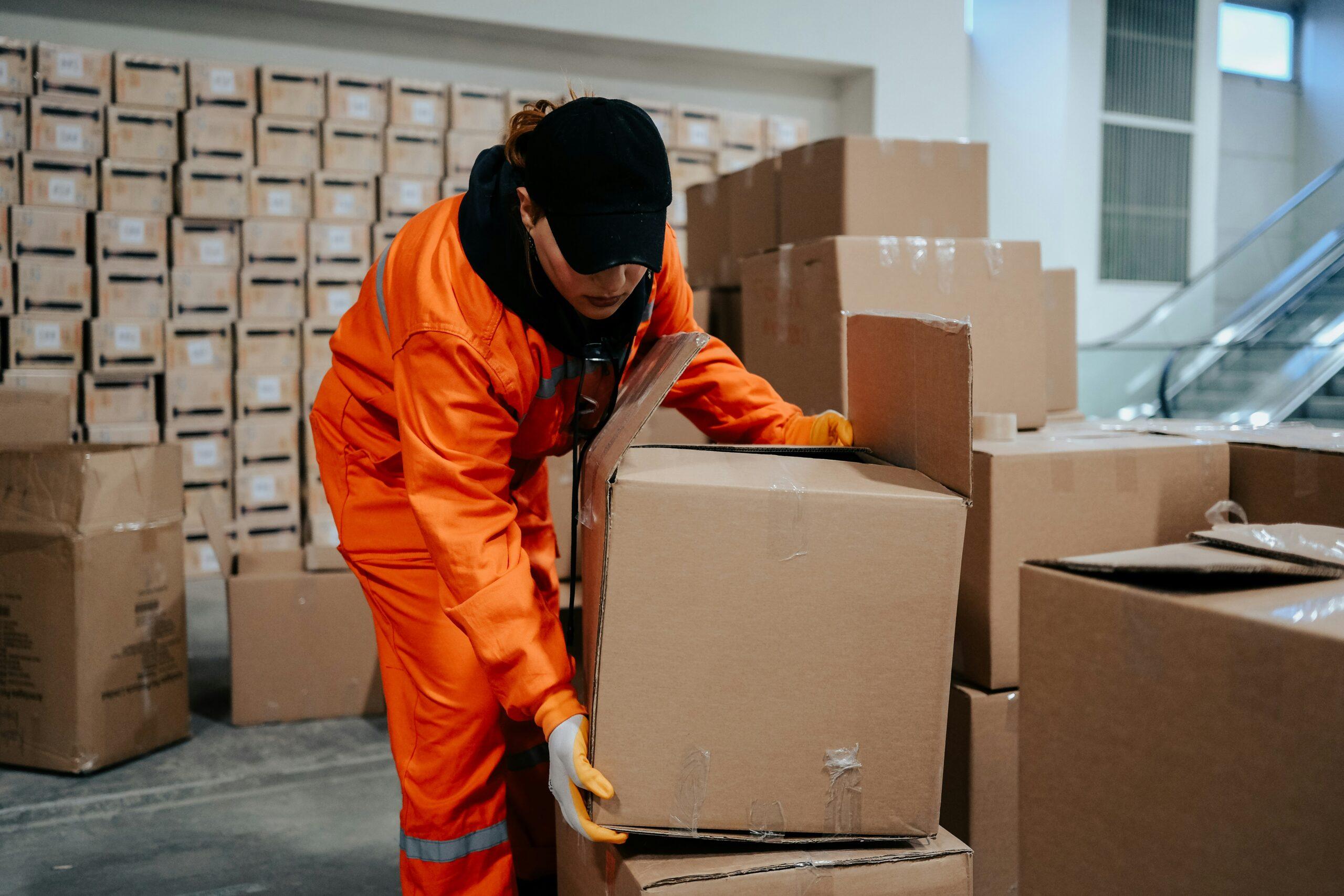 A worker in an orange safety uniform and black cap bending down to lift a large cardboard box in a warehouse filled with stacked packages.
