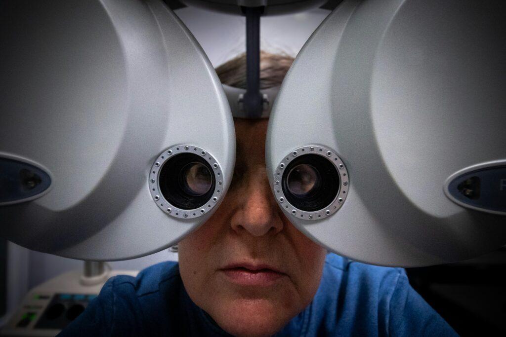 A person seated behind a large optometry phoropter during an eye exam, with their face framed by the two circular viewing lenses of the machine, surrounded by soft clinical lighting and equipment in the background.