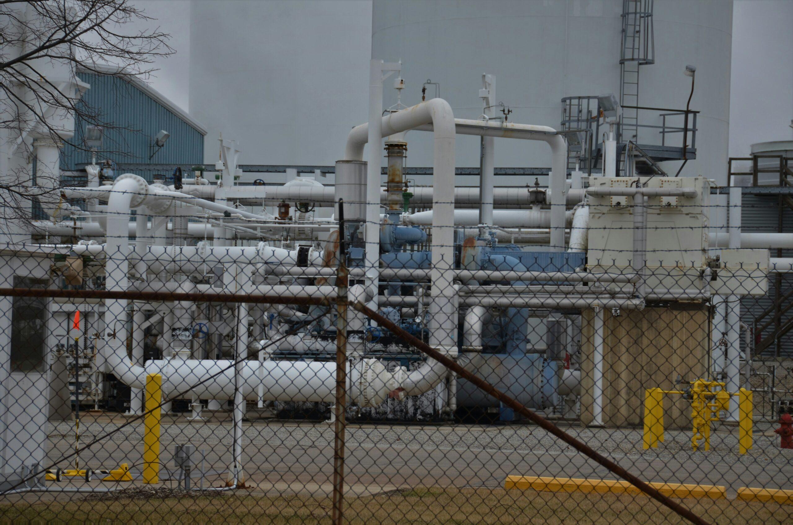 An industrial facility with large white storage tanks, interconnected metal pipes, valves, and processing equipment enclosed behind a chain-link fence, with yellow safety bollards and access platforms visible under an overcast sky.