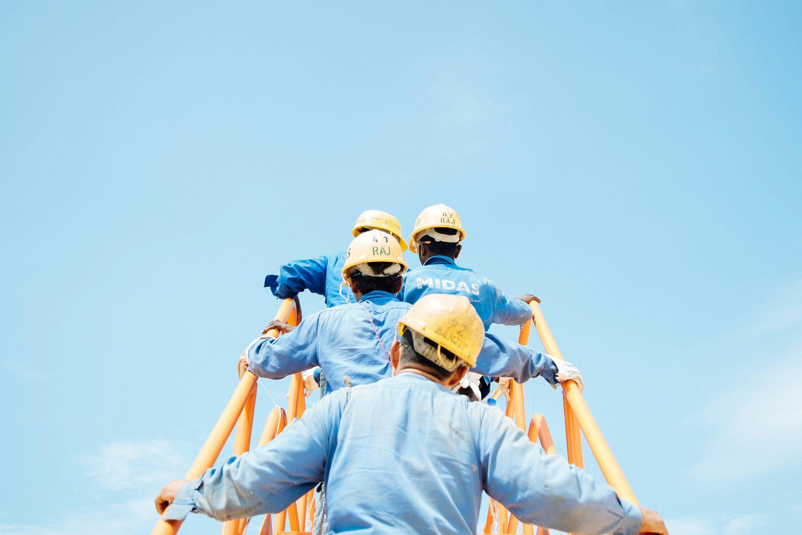 Workers in blue uniforms and yellow safety helmets climbing a tall orange industrial structure under a clear blue sky, viewed from behind as they ascend in a line.