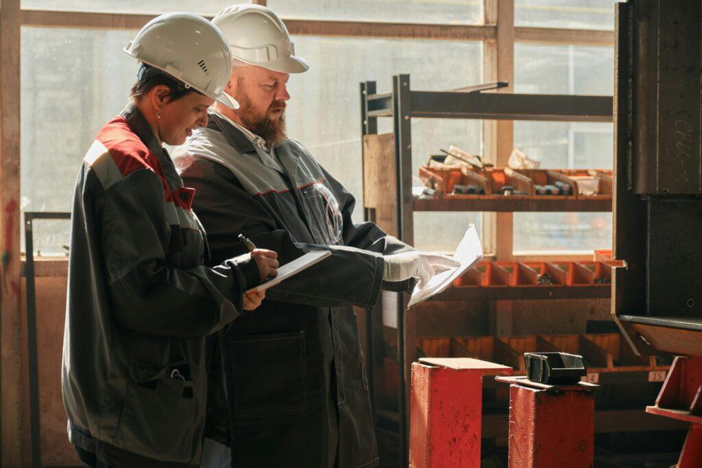 Man and woman wearing hard hats stand in a factory, surrounded by machinery and industrial equipment.