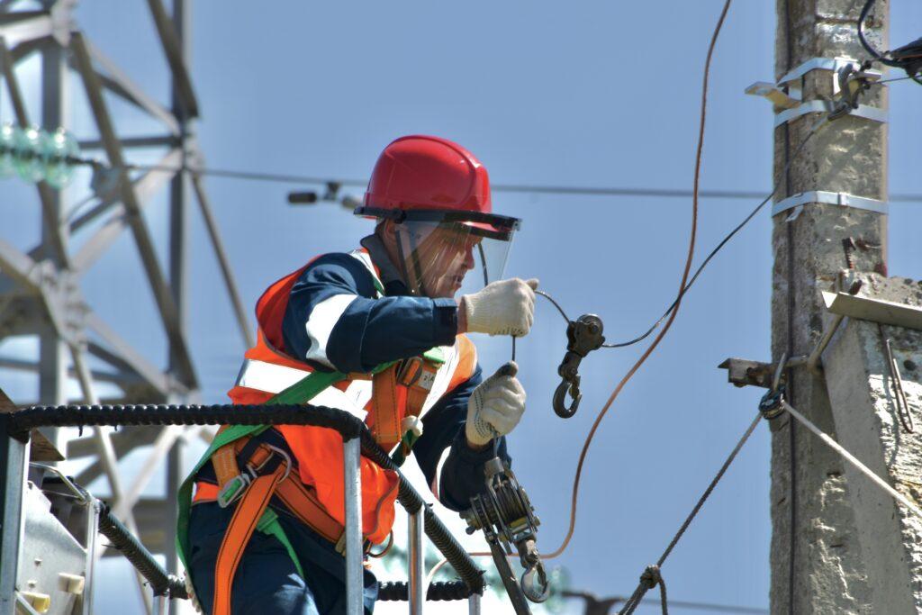 A man in an orange vest and safety glasses is working on an electrical pole, focused on his task.