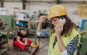 A female industrial supervisor wearing a yellow hard hat and high-visibility vest talks on a smartphone inside a factory, adjusting her helmet while looking concerned, with heavy machinery in the background and an injured worker sitting on the floor behind her.