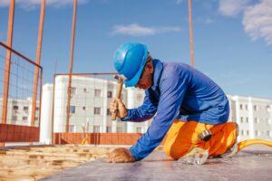 Construction worker installing roofing materials on a sloped roof under a clear blue sky.