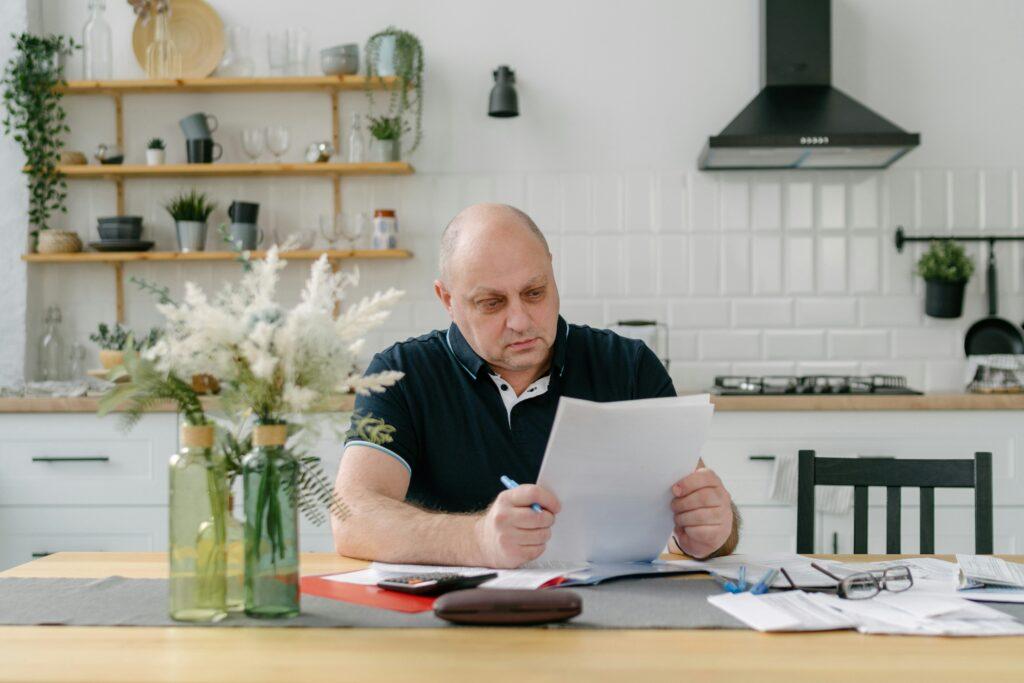 A bald man sits at a table with papers and a vase of flowers, focused on his work.