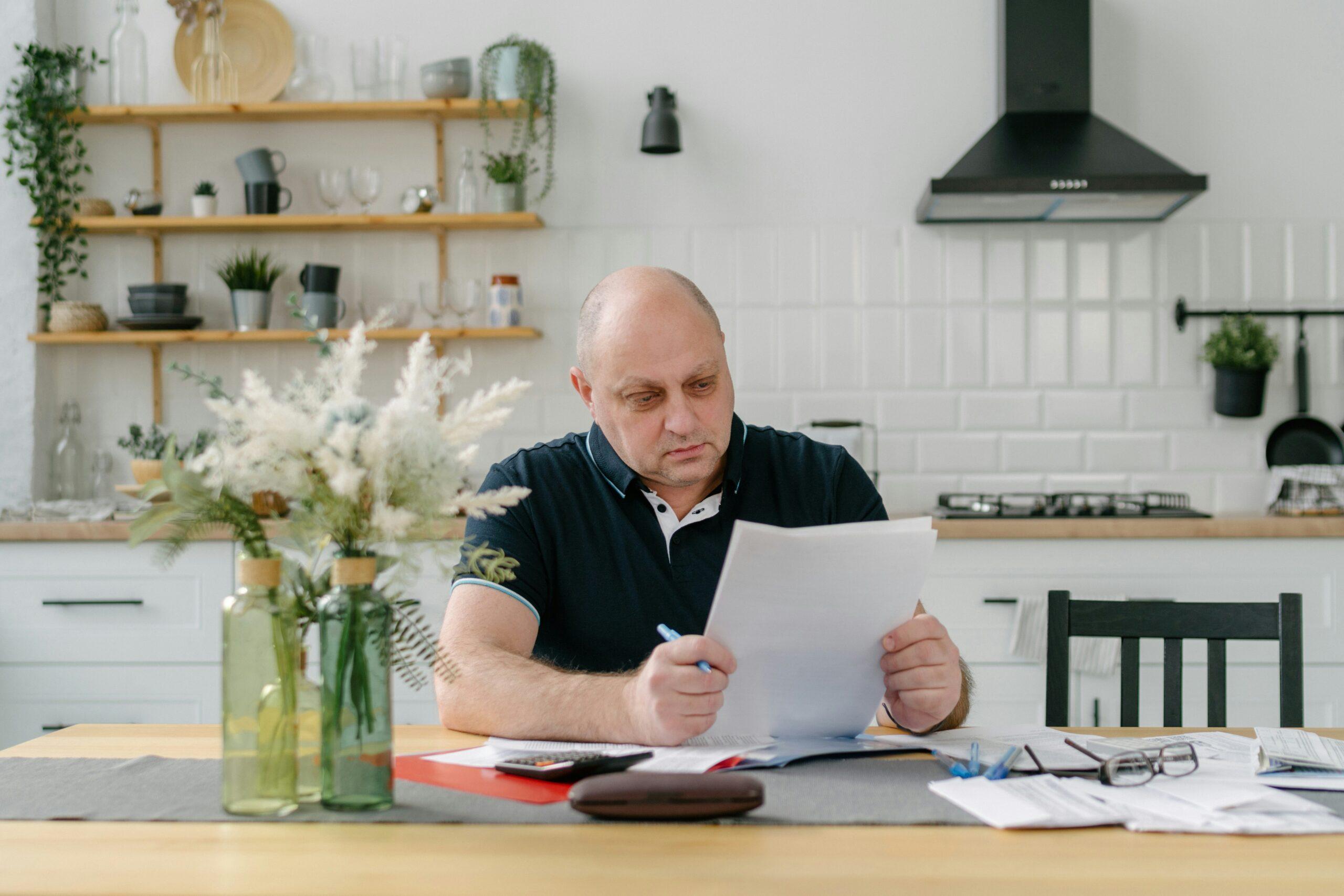A bald man sits at a table with papers and a vase of flowers, focused on his work.