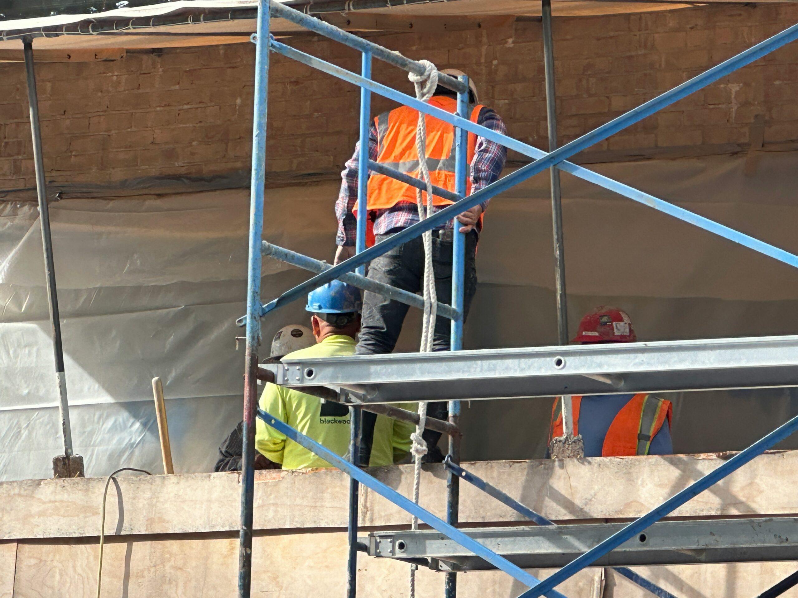 A construction worker wearing a hard hat stands on a scaffold, overseeing work on a building site.