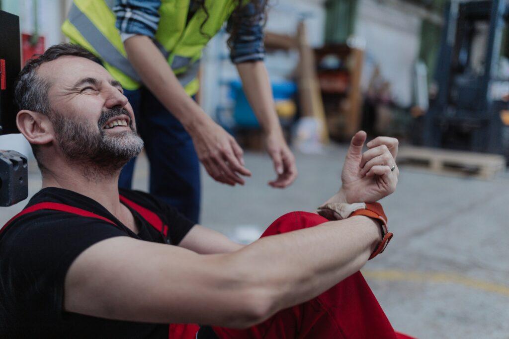 Injured worker sitting on the ground in a warehouse, grimacing in pain while holding his wrist as a coworker reaches out to help