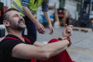 Injured worker sitting on the ground in a warehouse, grimacing in pain while holding his wrist as a coworker reaches out to help
