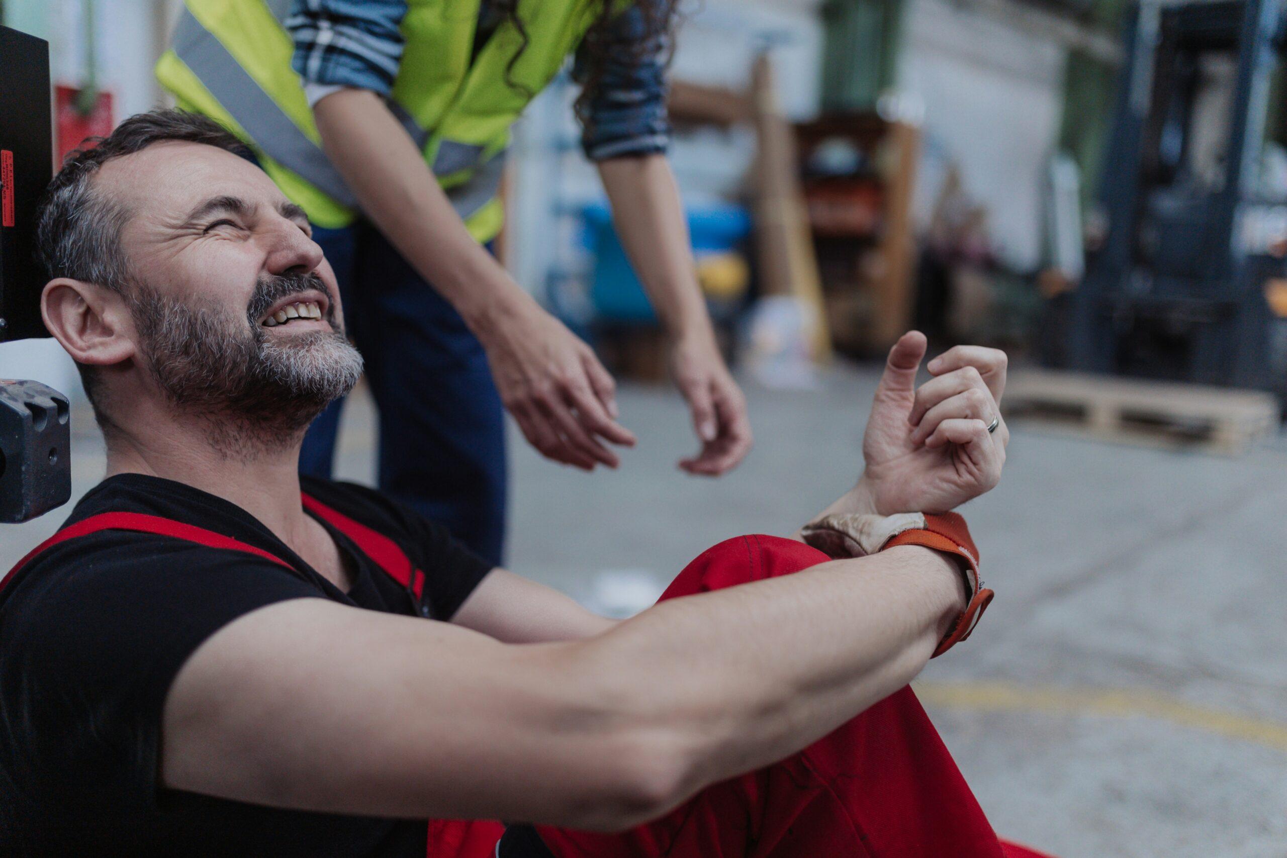 Injured worker sitting on the ground in a warehouse, grimacing in pain while holding his wrist as a coworker reaches out to help