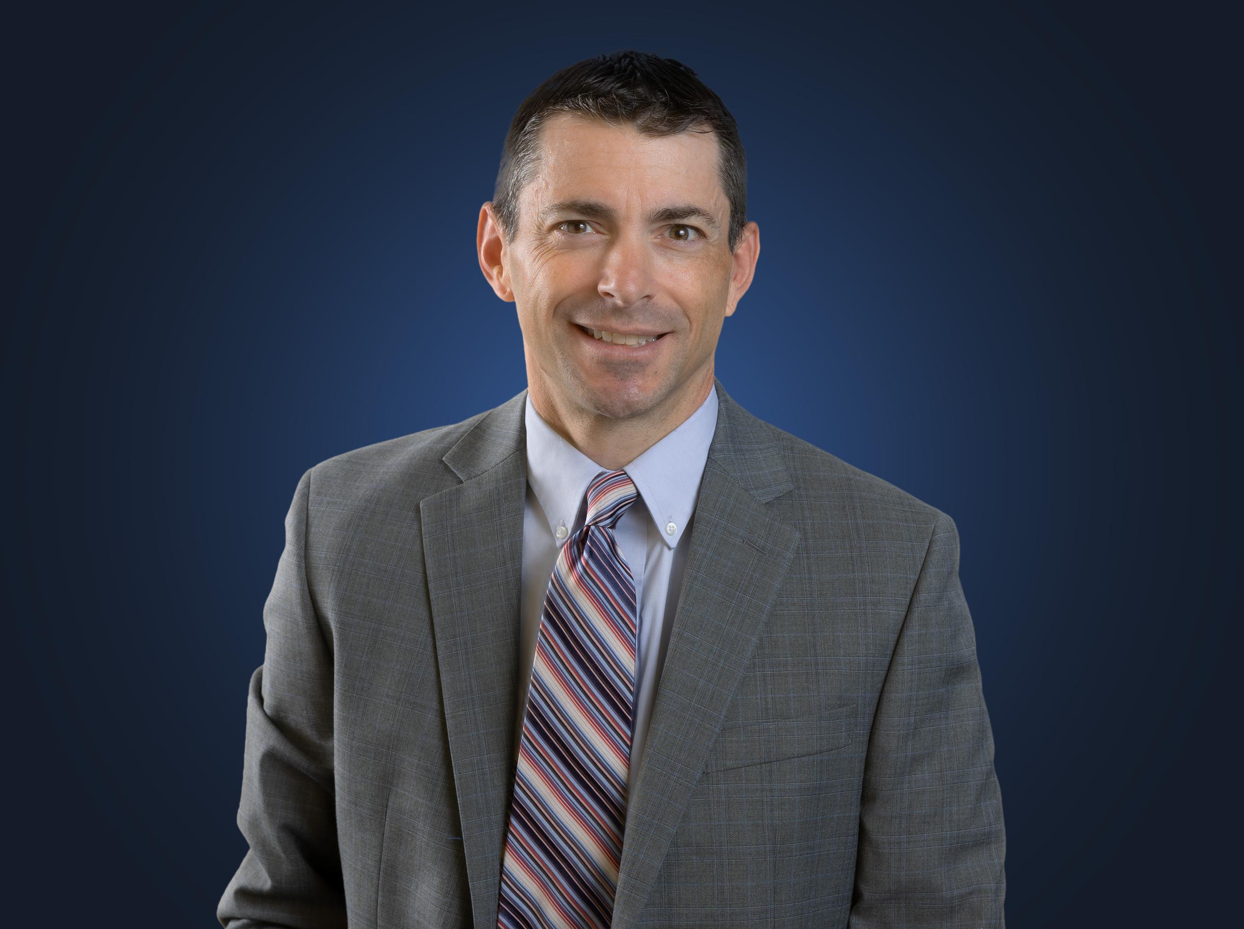 Attorney Lou D'Onofrio sits in front of a blue background while wearing a gray suit and striped tie