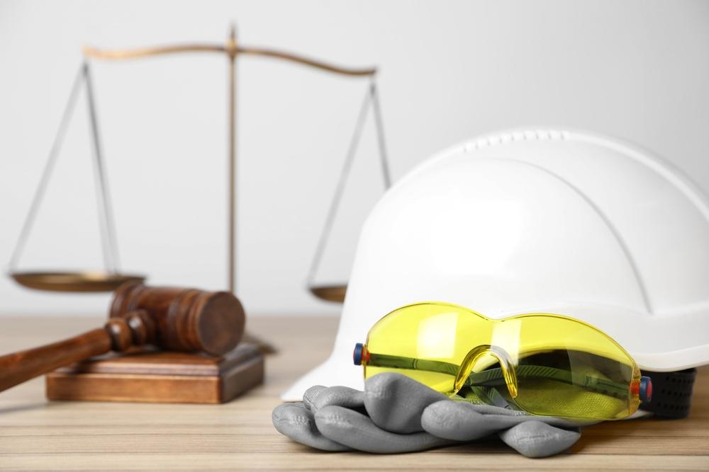 Hard hat, safety glasses, and work gloves on a desk with a gavel and scales of justice behind