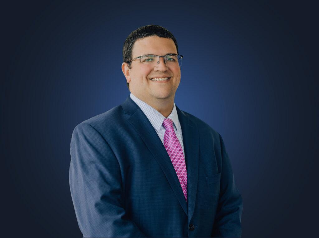 Attorney Brian Dunstone wears a dark suit and pink tie in front of a dark background