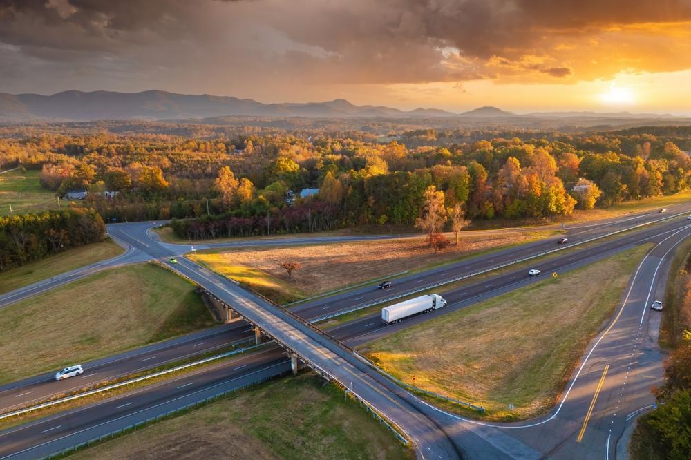 Aerial view of a highway interchange with cars and a semi-truck passing through a rural, wooded area at sunset